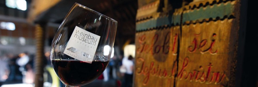 A wine glass with the logo of the Stuttgart Wine Museum is held in front of an old wooden barrel. People can be seen blurred in the background., © Stuttgart-Marketing GmbH, Thomas Niedermüller