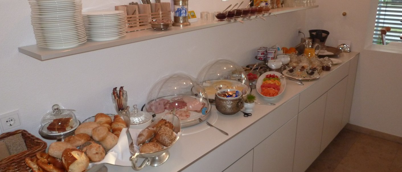 Ein Frühstücksbuffet mit Brötchen, Aufschnitt, Obst und Getränken auf einer weißen Theke in einem Hotel., © hotelmärchen GmbH Ein Frühstücksbuffet mit Brötchen, Aufschnitt, Obst und Getränken auf einer weißen Theke in einem Hotel., © hotelmärchen GmbH