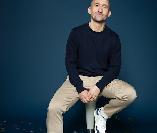 Person sitzt auf einem Hocker vor blauem Hintergrund, trägt dunklen Pullover und helle Hose. Goldene Konfetti liegen auf dem Boden., © Rosenau Kultur e.V. Person sitzt auf einem Hocker vor blauem Hintergrund, trägt dunklen Pullover und helle Hose. Goldene Konfetti liegen auf dem Boden., © Rosenau Kultur e.V.