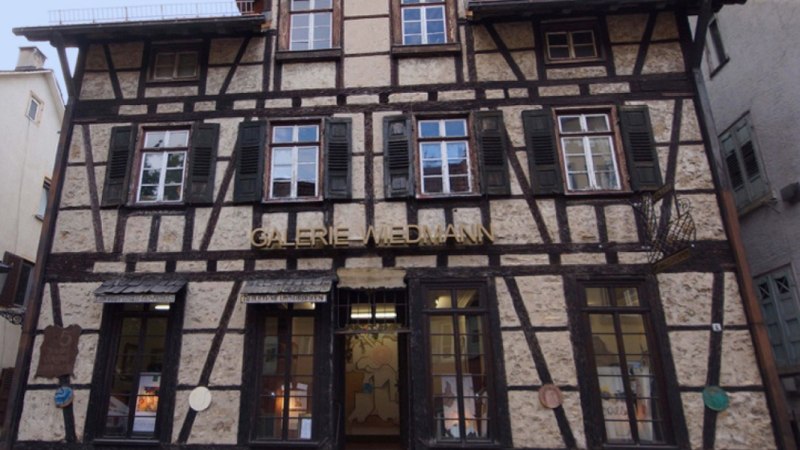 Fachwerkhaus mit der Aufschrift 'Galerie Wiedmann'. Das Gebäude hat mehrere Fenster mit grünen Fensterläden und eine Laterne an der Seite., © Galerie Wiedmann