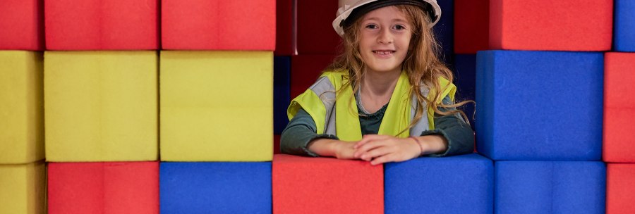 A child in a construction helmet and safety vest looks out smiling from a wall of colorful foam blocks. The blocks are red, yellow and blue., &copy; Julia Ochs