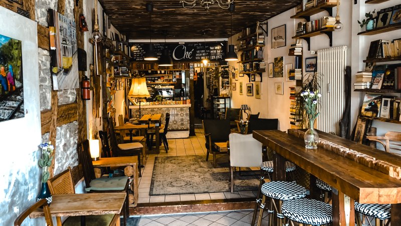 Ein gemütliches Café mit Holzmöbeln, Bücherregalen und Dekorationen an den Wänden. Einladende Atmosphäre mit warmem Licht., © Café Che Ein gemütliches Café mit Holzmöbeln, Bücherregalen und Dekorationen an den Wänden. Einladende Atmosphäre mit warmem Licht., © Café Che