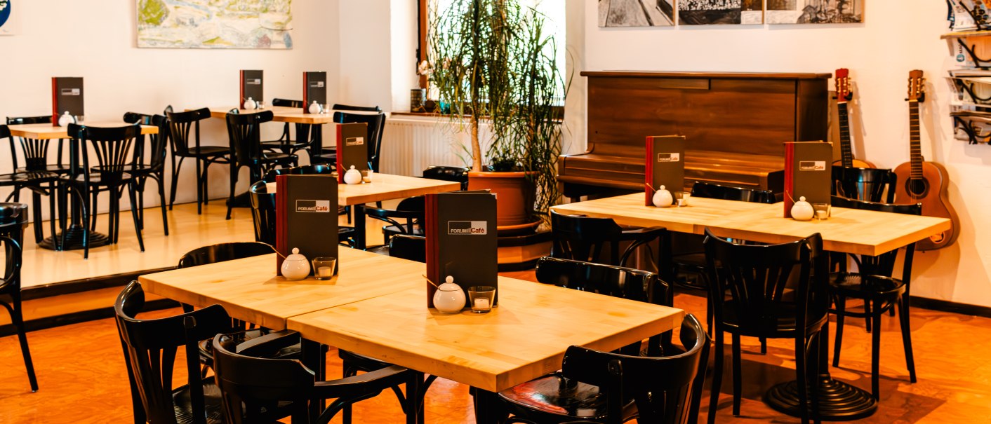 Gemütliches Café mit Holztischen, schwarzen Stühlen, einem Klavier und Gitarren an der Wand. Kunstwerke schmücken die Wände, Pflanzen sorgen für Atmosphäre., © SMG_Sarah Schmid