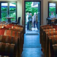 Barrique-Fässer in einer Vinothek, im Hintergrund Menschen und Tische mit Gläsern, durch eine offene Tür ist ein grüner Garten sichtbar., © Weingut Wöhrwag