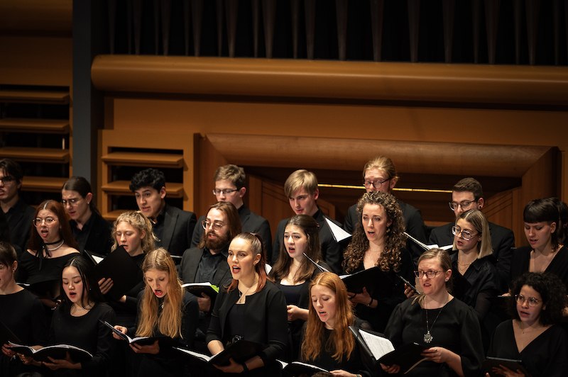 Ein Chor in schwarzer Kleidung singt in einem Konzertsaal. Die S&auml;ngerinnen und S&auml;nger halten Notenbl&auml;tter in den H&auml;nden., &copy; Staatliche Hochschule f&uuml;r Musik und Darstellende Kunst