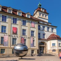 Das Linden-Museum in Stuttgart bei sonnigem Wetter. Vor dem Geb&auml;ude sind Plakate und eine gro&szlig;e silberne Kugel zu sehen. Menschen spazieren vorbei., &copy; Stuttgart-Marketing GmbH, Achim Mende