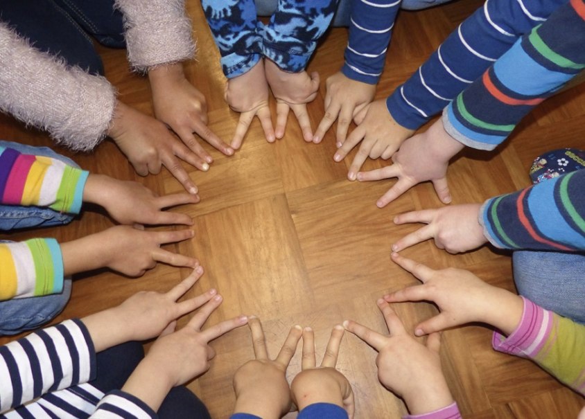 Kinderh&auml;nde in bunten &Auml;rmeln formen einen Kreis auf einem Holzboden. Die Finger zeigen zur Mitte und symbolisieren Gemeinschaft., &copy; Christian Steeneck