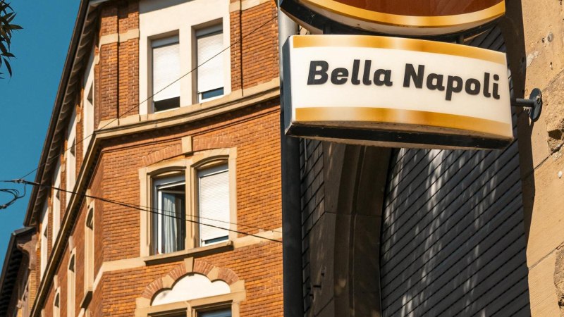 Ein Schild mit der Aufschrift 'Bella Napoli' hängt an einem Backsteingebäude. Der Himmel ist klar und blau, ein Baum ist im Vordergrund sichtbar., © SMG, Sarah Schmid Ein Schild mit der Aufschrift 'Bella Napoli' hängt an einem Backsteingebäude. Der Himmel ist klar und blau, ein Baum ist im Vordergrund sichtbar., © SMG, Sarah Schmid