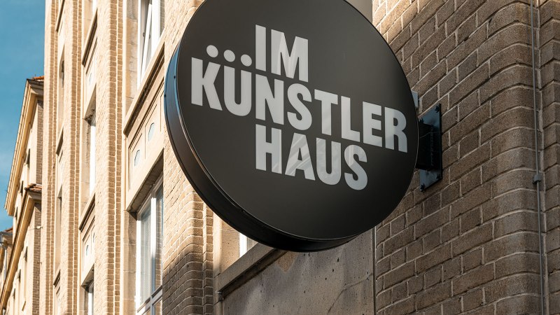 Rundes Schild mit 'IM KÜNSTLER HAUS' an einer Backsteinfassade, blauer Himmel im Hintergrund., © SMG, Sarah Schmid Rundes Schild mit 'IM KÜNSTLER HAUS' an einer Backsteinfassade, blauer Himmel im Hintergrund., © SMG, Sarah Schmid