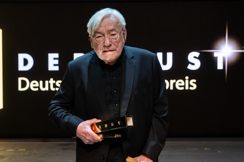 Ein älterer Mann in einem Anzug hält eine Trophäe vor einem Schild mit der Aufschrift 'DEUST Deutscher Preis'., © Württembergische Staatstheater Stuttgart