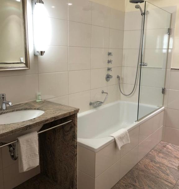 Modernes Badezimmer mit Badewanne, Duschkopf, Glasabtrennung und Waschbecken mit Spiegel. Helle Fliesen und Handtuchhalter sind sichtbar., © Hotel Royal