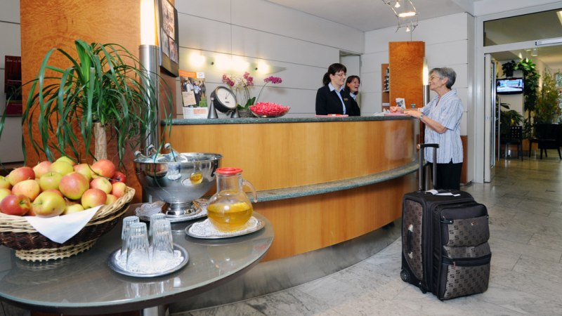 A hotel reception desk with two employees helping an elderly woman with a suitcase. A table with apples and drinks is in the foreground., &copy; Hotel Unger