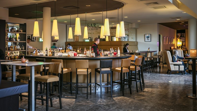Modern bar with round counter, hanging lamps and seating areas in the Arcotel Camino. People sit and enjoy the atmosphere., © © Harald Eisenberger Modern bar with round counter, hanging lamps and seating areas in the Arcotel Camino. People sit and enjoy the atmosphere., © © Harald Eisenberger