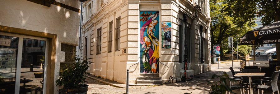 Eine Stra&szlig;enecke mit einem Pub-Schild, bunter Wandkunst und Caf&eacute;-Tischen unter einem Sonnenschirm., &copy; SMG Stuttgart Marketing GmbH - Sarah Schmid
