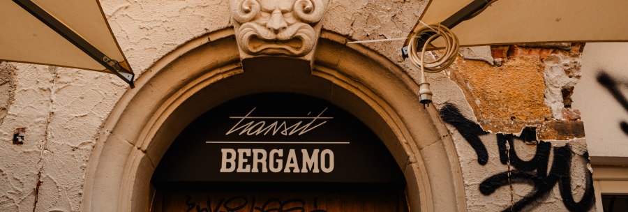 Stone relief of a child above an entrance with the inscription 'transit Bergamo'. The plaster is crumbling, graffiti and awnings are visible., © Sarah Schmid Stone relief of a child above an entrance with the inscription 'transit Bergamo'. The plaster is crumbling, graffiti and awnings are visible., © Sarah Schmid
