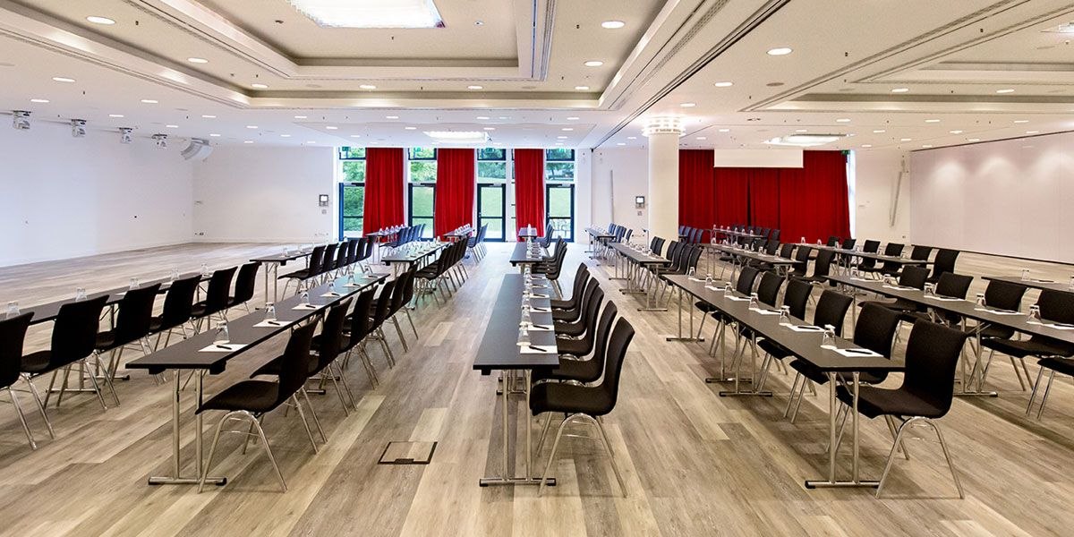 A large conference room with long tables, black chairs, red curtains, and bright lighting., © Dormero Hotel Stuttgart A large conference room with long tables, black chairs, red curtains, and bright lighting., © Dormero Hotel Stuttgart