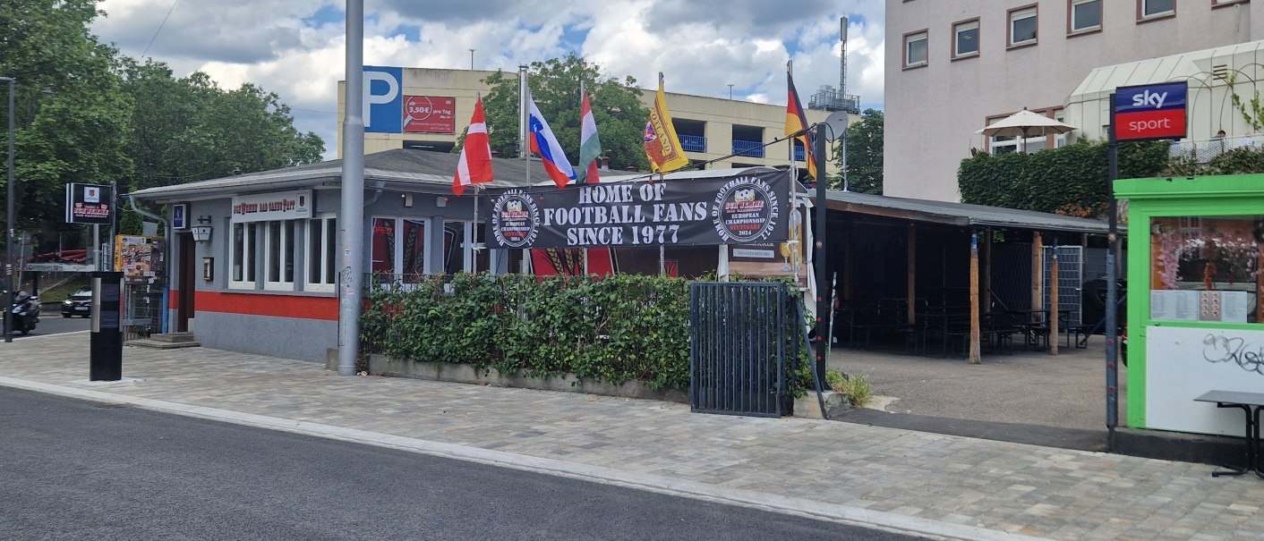 Bar mit Fahnen und Schild "Home of Football Fans Since 1977". Gebäude mit roten und grauen Akzenten, umgeben von Bäumen und einem Parkplatz., © Stuttgart-Marketing GmbH Bar mit Fahnen und Schild "Home of Football Fans Since 1977". Gebäude mit roten und grauen Akzenten, umgeben von Bäumen und einem Parkplatz., © Stuttgart-Marketing GmbH