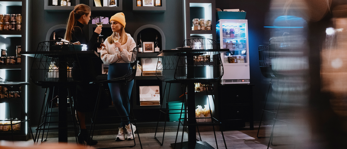 Zwei Frauen in einem stilvollen Café, eine mit gelber Mütze, halten Weingläser. Im Hintergrund sind Regale mit Produkten und ein Kühlschrank zu sehen., © Stuttgart-Marketing GmbH, Alwin Maigler