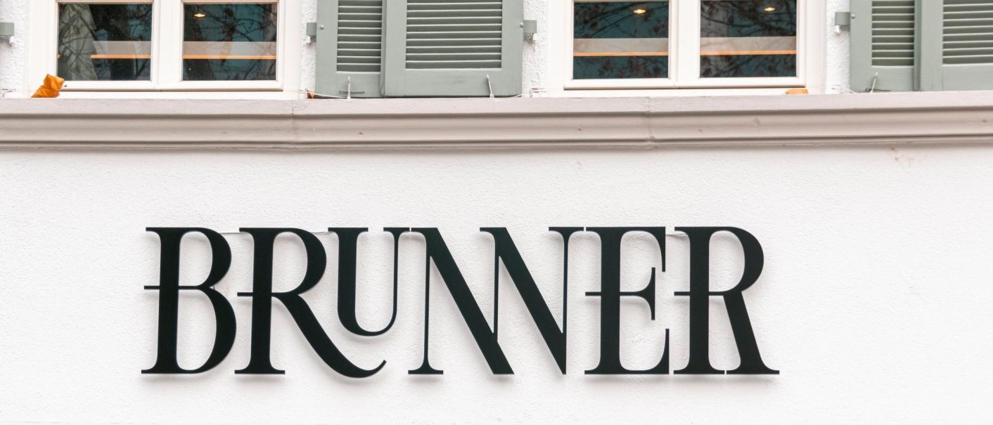Schriftzug an der Hausfassade des Brunner Restaurants mit Serifen. Darüber Sprossenfenster mit grünen Fensterläden., © SMG, Sarah Schmid Schriftzug an der Hausfassade des Brunner Restaurants mit Serifen. Darüber Sprossenfenster mit grünen Fensterläden., © SMG, Sarah Schmid