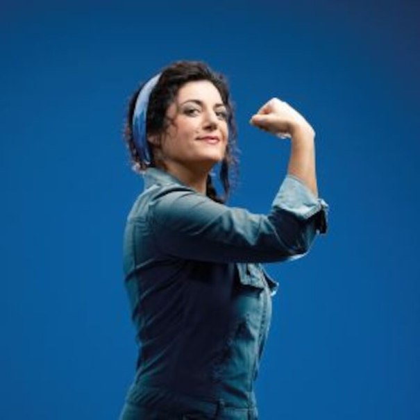 Woman in work clothes with headscarf shows a strong pose against a blue background, inspired by Rosie the Riveter., © Altes Schauspielhaus und Komödie im Marquardt e.V. Woman in work clothes with headscarf shows a strong pose against a blue background, inspired by Rosie the Riveter., © Altes Schauspielhaus und Komödie im Marquardt e.V.