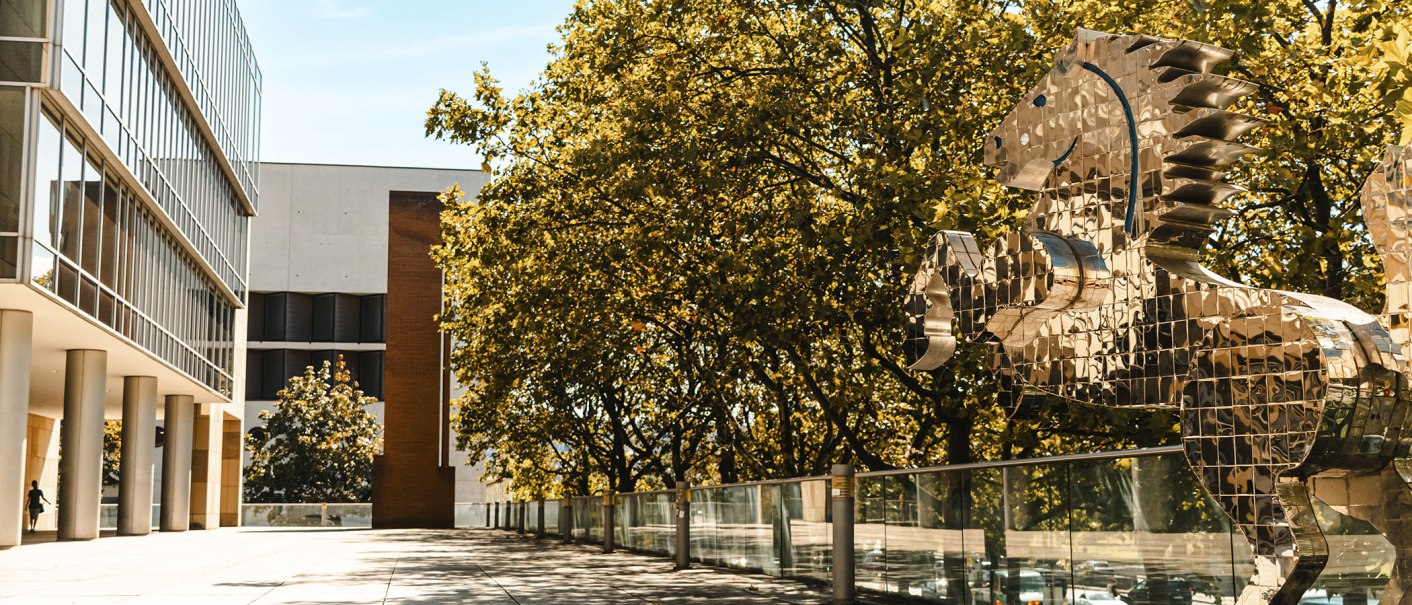 Moderne Glasfassade neben einer glänzenden Pferdeskulptur aus Spiegeln, umgeben von Bäumen und blauem Himmel., © Sarah Schmid Moderne Glasfassade neben einer glänzenden Pferdeskulptur aus Spiegeln, umgeben von Bäumen und blauem Himmel., © Sarah Schmid