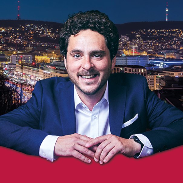 Ein Mann in einem Anzug lächelt vor einer beleuchteten Stadt bei Nacht. Im Hintergrund sind Lichter und Gebäude zu sehen., © Altes Schauspielhaus und Komödie im Marquardt e.V.