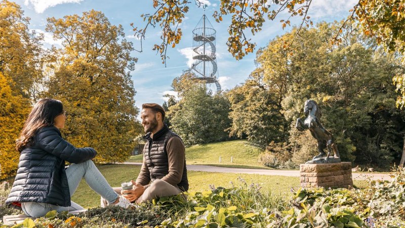 Zwei Personen sitzen im Höhenpark Killesberg, umgeben von Herbstbäumen. Im Hintergrund ist der Killesbergturm zu sehen, neben einer Pferdestatue., © Stuttgart-Marketing GmbH, Sarah Schmid