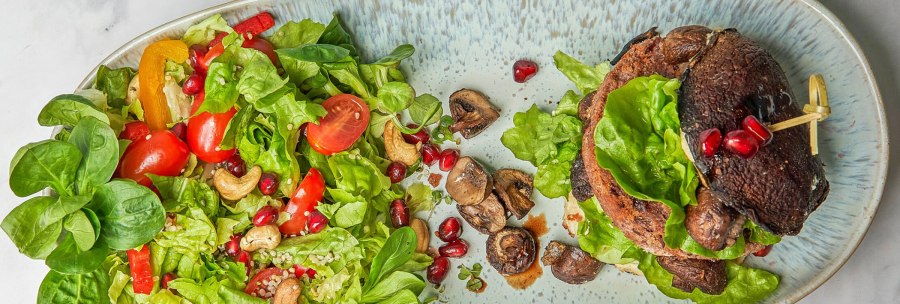 A plate with a salad of green leafy vegetables, tomatoes, peppers, cashew nuts and pomegranate seeds. Next to it, a burger with mushrooms., © SMG A plate with a salad of green leafy vegetables, tomatoes, peppers, cashew nuts and pomegranate seeds. Next to it, a burger with mushrooms., © SMG