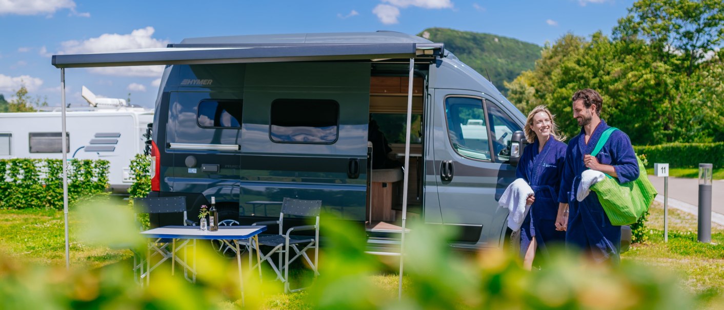 A couple in blue bathrobes is standing, smiling, in front of a modern RV at a sunny campground., © Thomas Niedermüller A couple in blue bathrobes is standing, smiling, in front of a modern RV at a sunny campground., © Thomas Niedermüller