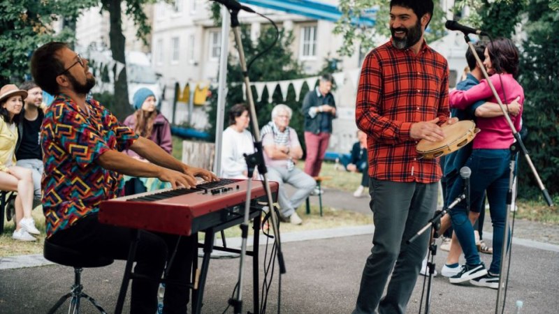 Zwei Musiker spielen im Freien. Einer sitzt am Keyboard, der andere steht mit einem Tamburin. Zuschauer sitzen im Hintergrund., © Nico Frank Zwei Musiker spielen im Freien. Einer sitzt am Keyboard, der andere steht mit einem Tamburin. Zuschauer sitzen im Hintergrund., © Nico Frank