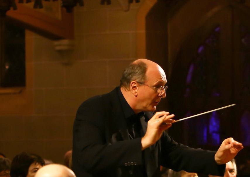 Ein Dirigent in einem Konzertsaal dirigiert mit einem Taktstock. Im Hintergrund sind unscharfe Zuschauer zu sehen., © KCBW