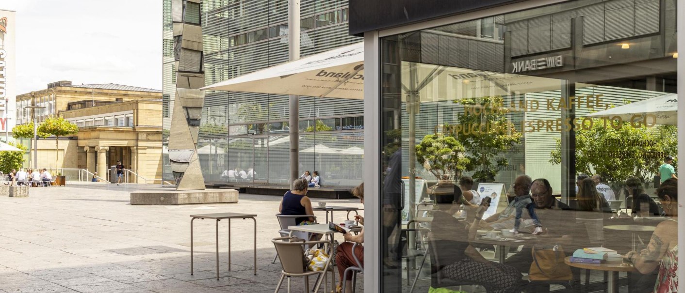 Menschen sitzen in einem Café auf einem städtischen Platz. Moderne Gebäude und ein Kunstwerk sind im Hintergrund zu sehen., © SMG, Sarah Schmid