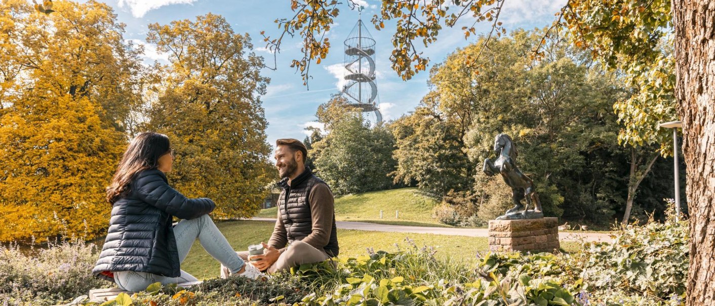 Zwei Personen sitzen im Höhenpark Killesberg, umgeben von Herbstbäumen. Im Hintergrund ist der Killesbergturm zu sehen, neben einer Pferdestatue., © Stuttgart-Marketing GmbH, Sarah Schmid Zwei Personen sitzen im Höhenpark Killesberg, umgeben von Herbstbäumen. Im Hintergrund ist der Killesbergturm zu sehen, neben einer Pferdestatue., © Stuttgart-Marketing GmbH, Sarah Schmid