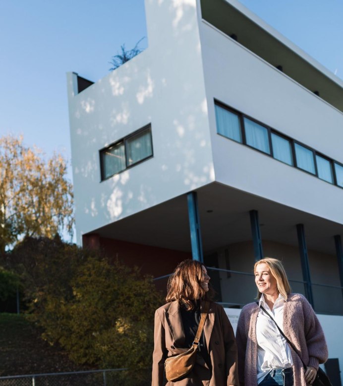 Zwei Frauen spazieren vor einem modernen, weißen Gebäude der Weißenhofsiedlung in Stuttgart. Der Himmel ist klar und blau., © SMG, wpsteinheisser Zwei Frauen spazieren vor einem modernen, weißen Gebäude der Weißenhofsiedlung in Stuttgart. Der Himmel ist klar und blau., © SMG, wpsteinheisser