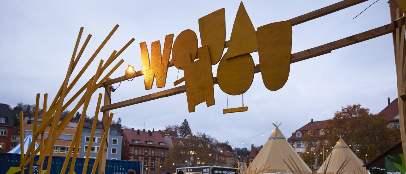 Entrance to the Wouahou Winter Village with yellow sign and tepees in the background. Buildings and trees surround the square., © SMG, Sarah Schmid Entrance to the Wouahou Winter Village with yellow sign and tepees in the background. Buildings and trees surround the square., © SMG, Sarah Schmid