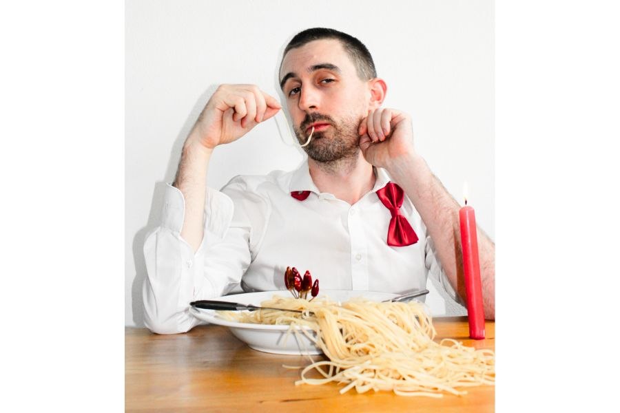 Ein Mann in wei&szlig;em Hemd mit roter Fliege isst l&auml;ssig Spaghetti. Eine rote Kerze steht auf dem Tisch und sorgt f&uuml;r eine humorvolle Atmosph&auml;re., &copy; Fotocredit Theater La Lune Stuttgart
