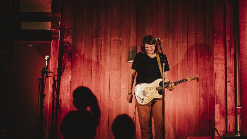 Ein Musiker steht l&auml;chelnd mit einer E-Gitarre auf einer B&uuml;hne vor einer Holzwand, beleuchtet von warmem, rotem Licht., &copy; Stefan Ebert