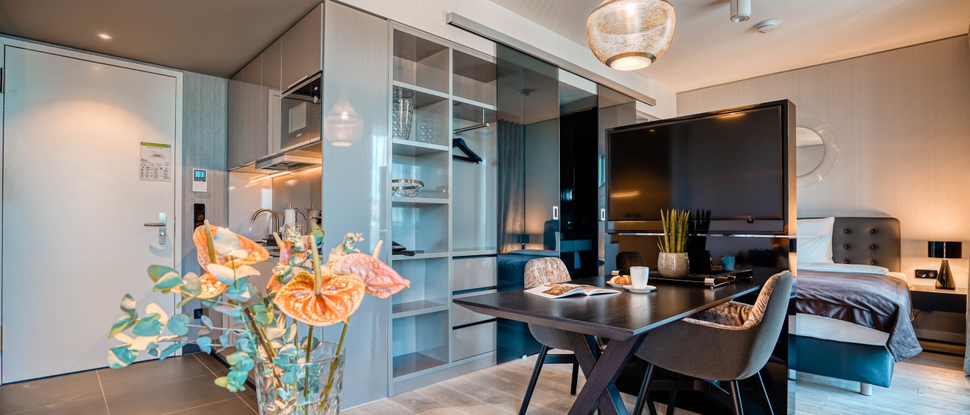 Moderne Suite mit Esstisch, Bett, kleiner Küche und stilvoller Dekoration. Ein großer Fernseher trennt den Schlafbereich vom Wohnbereich., © CLOUD N°7 APARTMENTS GmbH Moderne Suite mit Esstisch, Bett, kleiner Küche und stilvoller Dekoration. Ein großer Fernseher trennt den Schlafbereich vom Wohnbereich., © CLOUD N°7 APARTMENTS GmbH