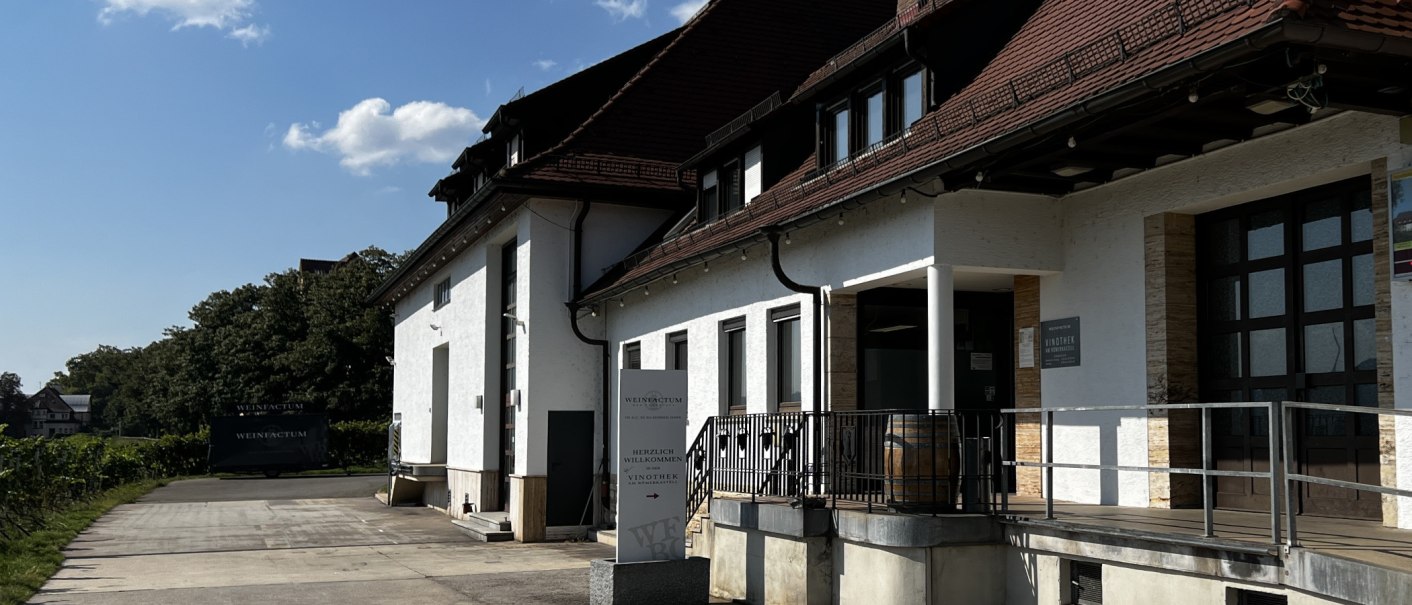 Weißes Gebäude mit rotem Dach, Vinothek-Schild und Weinfass vor dem Eingang. Umgeben von Bäumen und klarem Himmel., © Weinfactum Bad Cannstatt