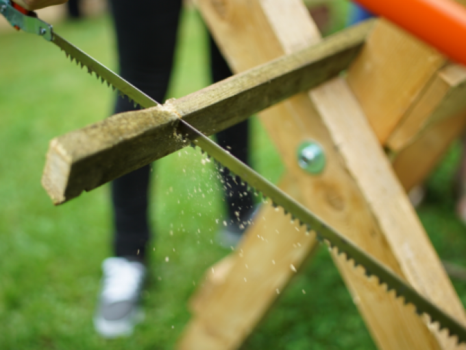 Eine Person sägt mit einer Handsäge ein Holzstück auf einer Holzhalterung im Freien. Späne fliegen umher., © Cool-Tours StattReisen Eine Person sägt mit einer Handsäge ein Holzstück auf einer Holzhalterung im Freien. Späne fliegen umher., © Cool-Tours StattReisen