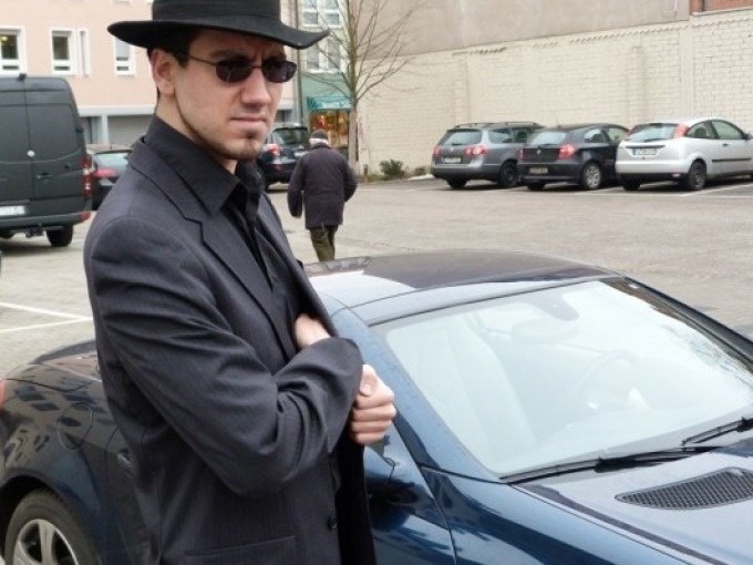 Ein Mann in dunklem Anzug und Hut steht neben einem blauen Auto auf einem Parkplatz. Im Hintergrund sind weitere Autos und Geb&auml;ude zu sehen., &copy; Cool-Tours StattReisen