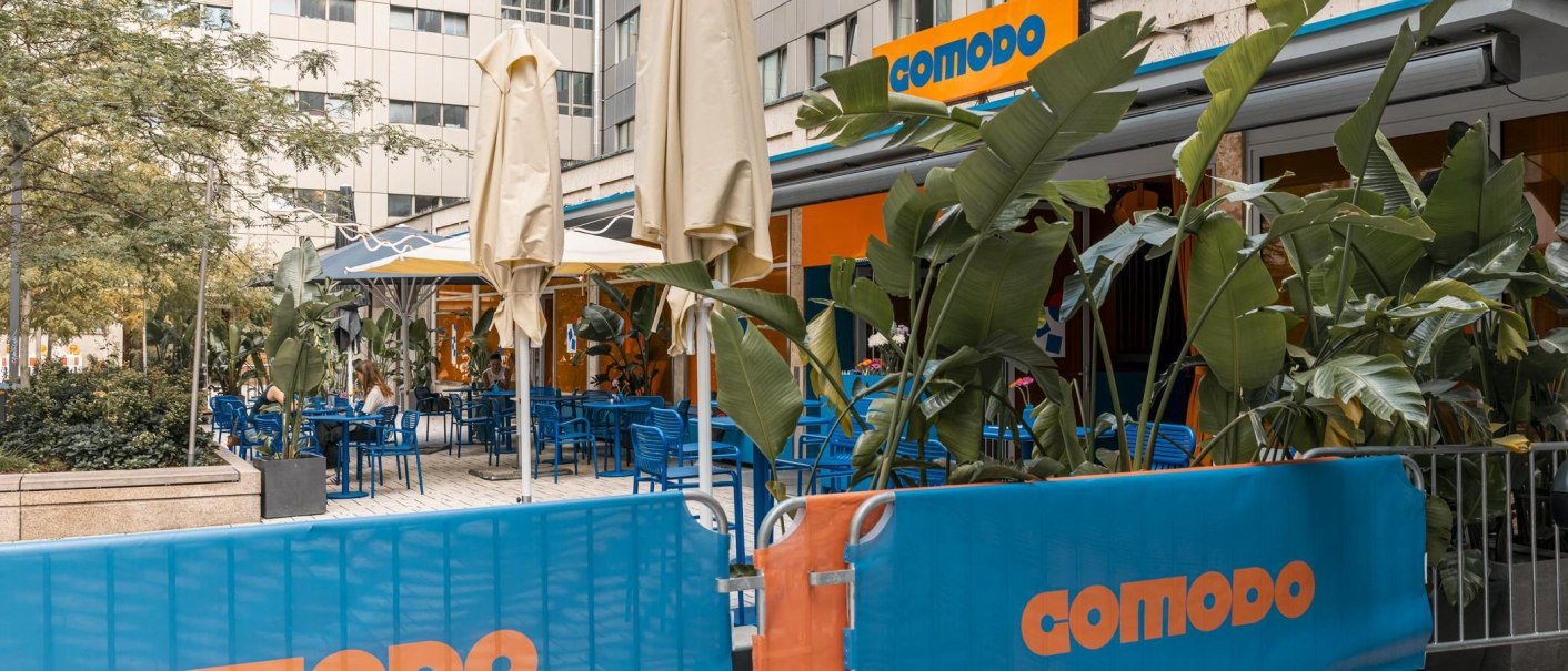 Outdoor area of a caf&eacute; with blue chairs, tables and plants. The 'comodo' sign is visible. Parasols are set up., &copy; Stuttgart Marketing GmbH, Sarah Schmid