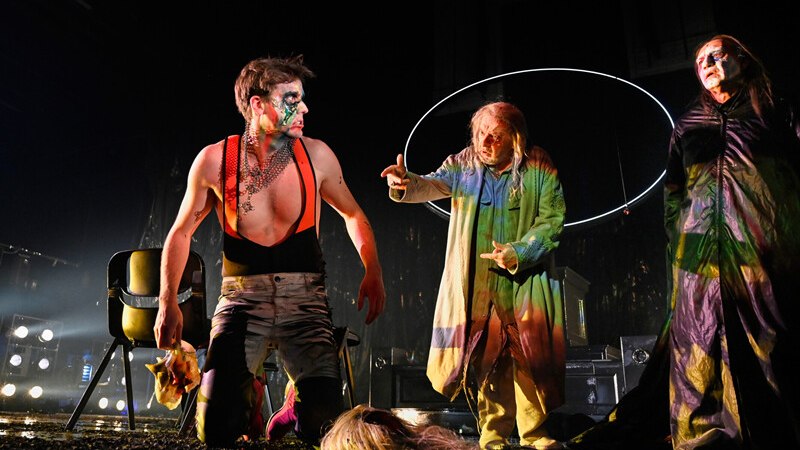 Drei Schauspieler in farbenfrohen Kostümen auf einer Bühne. Einer kniet, während die anderen stehen. Die Szene ist dramatisch beleuchtet., © Württembergische Staatstheater Stuttgart