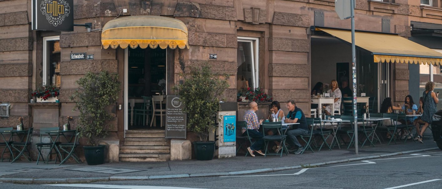 Café mit gelbem Vordach an einer Straßenecke. Gäste sitzen draußen an Tischen. Schild mit 'Lumen' und Straßenschild 'Schwabstr.' sichtbar., © SMG, Sarah Schmid
