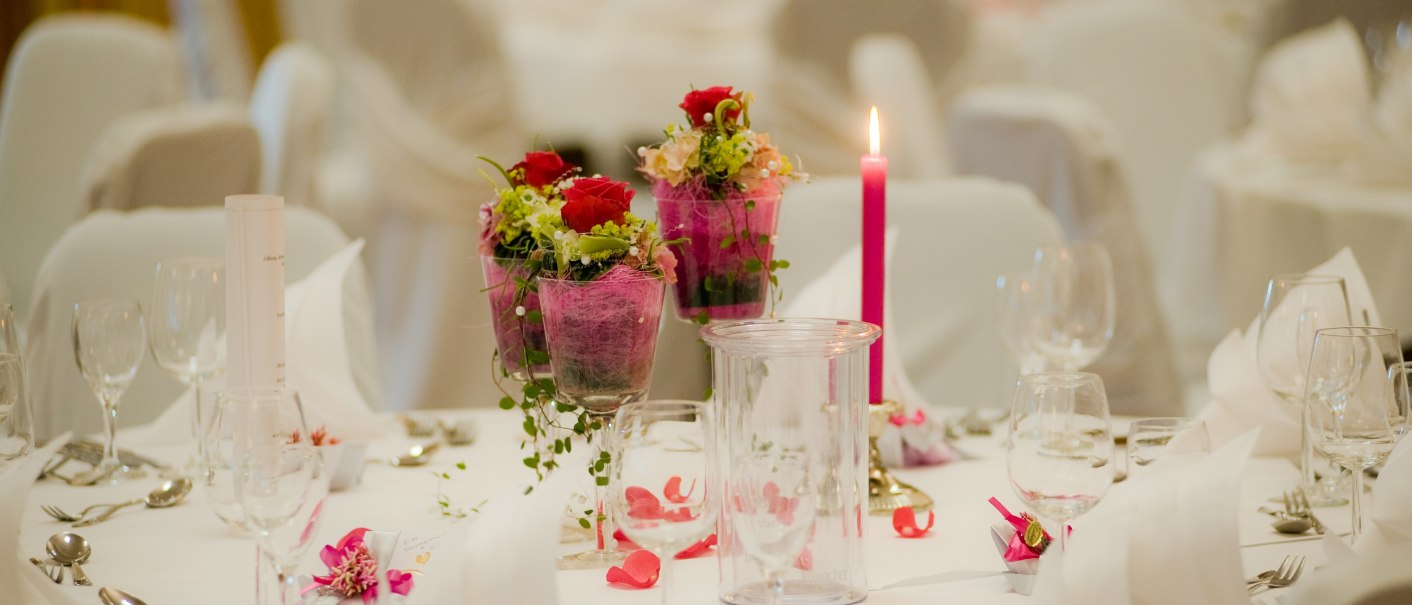 Festlich gedeckter Tisch mit weißen Tischdecken, roten Blumenarrangements und einer brennenden rosa Kerze als Dekoration., © Hotel Fortuna