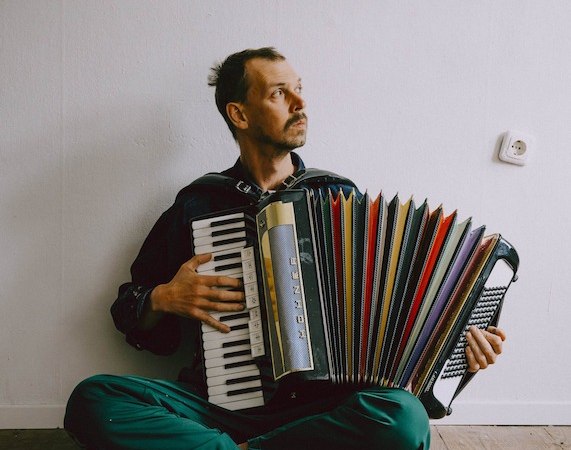 A man sits on the wooden floor and plays an accordion. He wears green pants and looks thoughtfully to the side., &copy; Rosenau Kultur e.V.