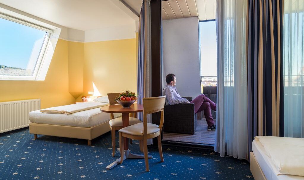 Hotelzimmer mit zwei Betten, Tisch und St&uuml;hlen. Ein Mann sitzt auf dem Balkon. Helle W&auml;nde und Teppichboden mit Muster., &copy; Aparthotel Astro