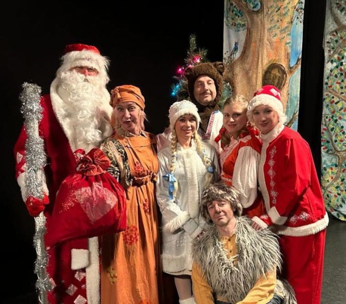 Menschen in festlichen Kostümen, darunter Weihnachtsmann und traditionelle russische Kleidung, posieren vor einem bemalten Hintergrund mit Baum und Vogel., © Theater Atelier