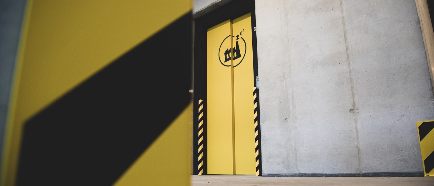 Gelbe Tür mit schwarzem Logo und Zzz-Symbol in einem Betonraum. Die Tür hat schwarz-gelbe Streifen an den Seiten., © Das Schlafwerk - DQuadrat Living GmbH