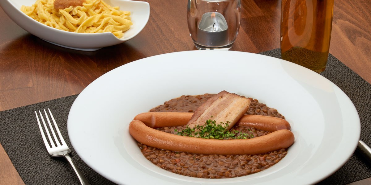 Linseneintopf mit Würstchen und Speck in einem weißen Teller, daneben eine Schüssel Spätzle auf einem Holztisch., © Stuttgart-Marketing GmbH Linseneintopf mit Würstchen und Speck in einem weißen Teller, daneben eine Schüssel Spätzle auf einem Holztisch., © Stuttgart-Marketing GmbH